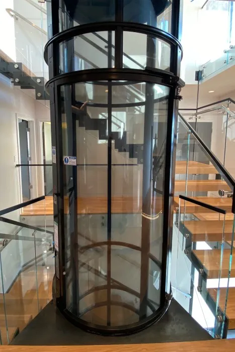 Round PVE home elevator with black powder coated frame beside a staircase with glass railings