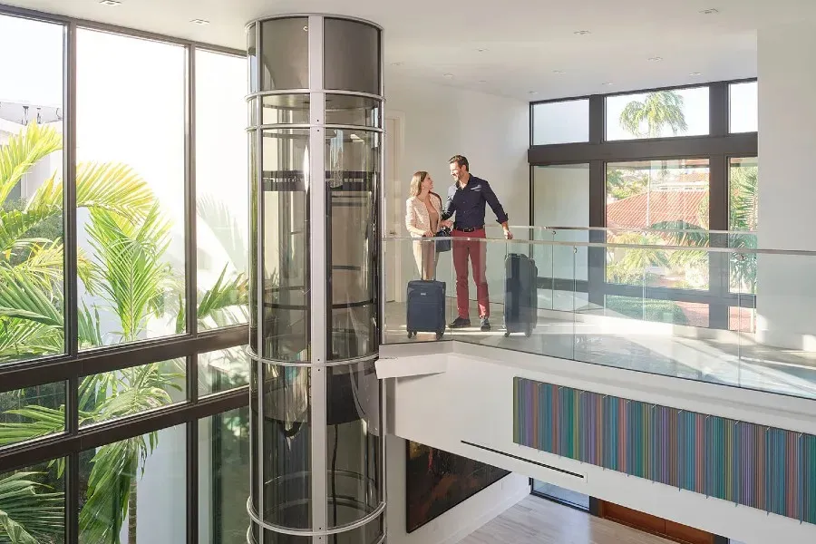 Couple with luggage beside a cylindrical PVE home elevator with automatic homing feature on an upper floor landing