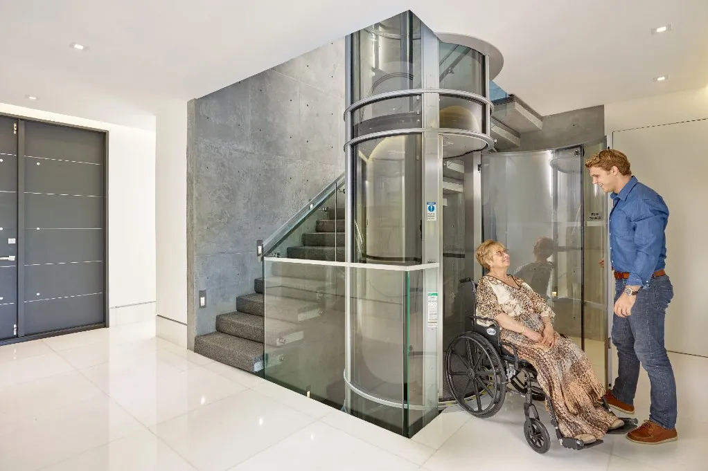 Woman in a wheelchair beside a glass PVE home elevator with a man helping her near a staircase