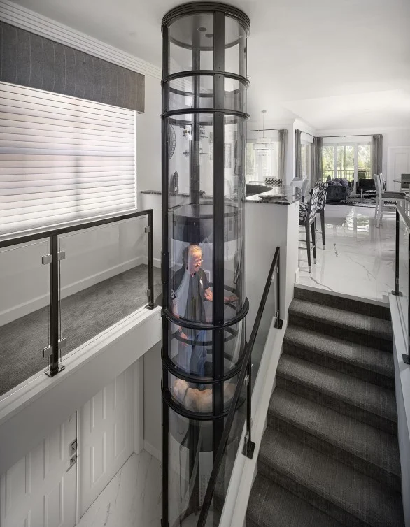 Person riding a two stop PVE pneumatic vacuum elevator with glass tube beside a staircase in an open plan home