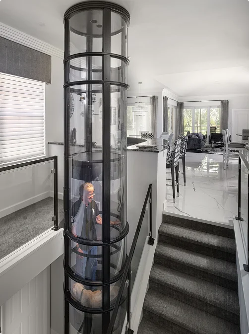 Person riding a PVE pneumatic vacuum elevator in a glass tube beside a staircase in an open concept home