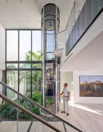 Woman rides a PVE home elevator in a glass tube as a man waits in a bright modern foyer